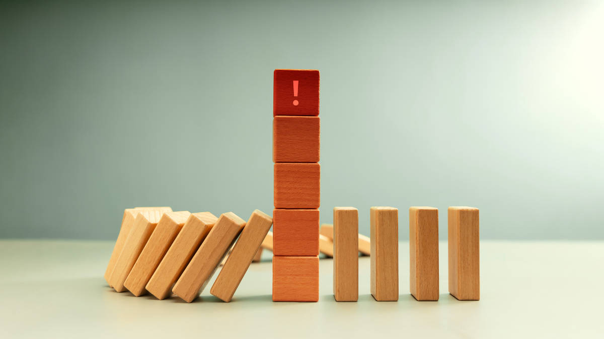Rotes Warnschild auf einem Holzklotzturm inmitten fallender Dominosteine, symbolisch für Risiko- und Krisenmanagement.