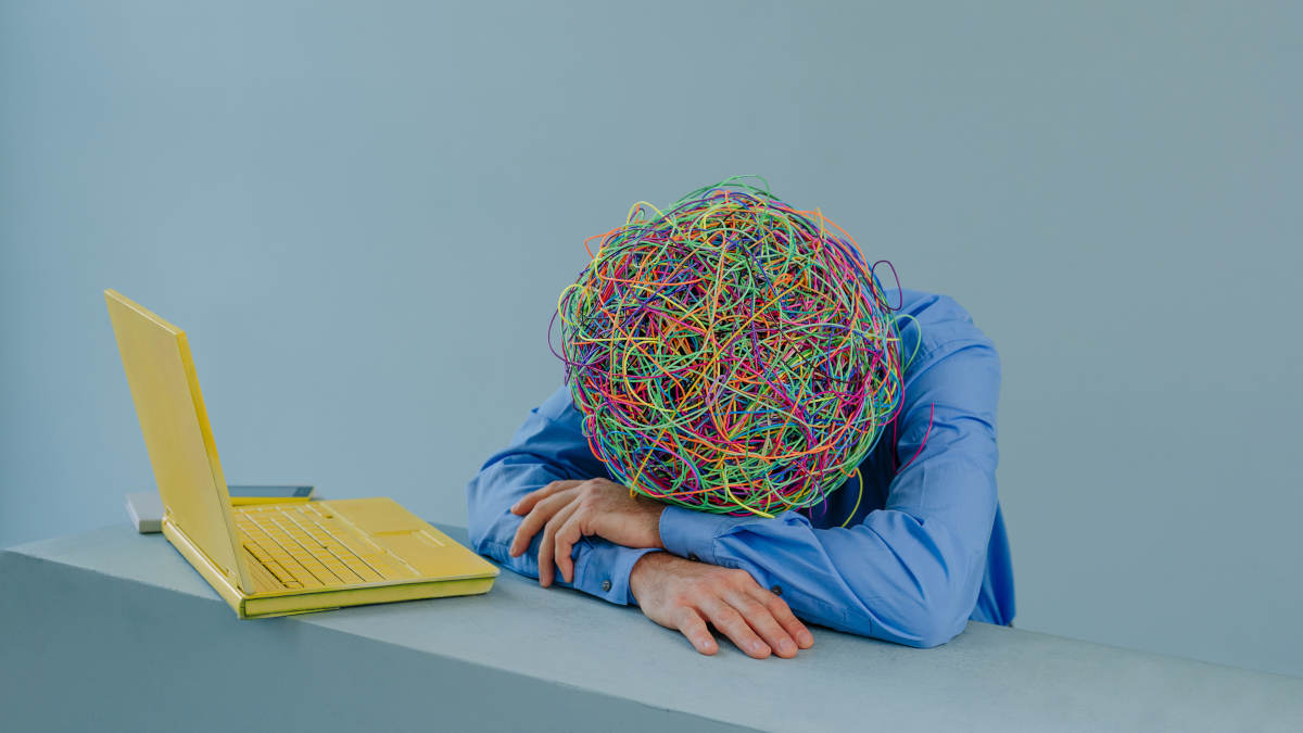 Person mit Gummiband-Knäuel statt Kopf liegt vor gelbem Laptop auf Tisch.