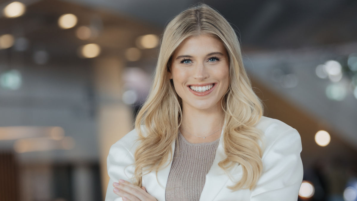 Portrait Foto der Unternehmerin Céline Flores Willers. Sie trägt einen weißen Blazer und verschränkt die Arme und lächelt in die Kamera.