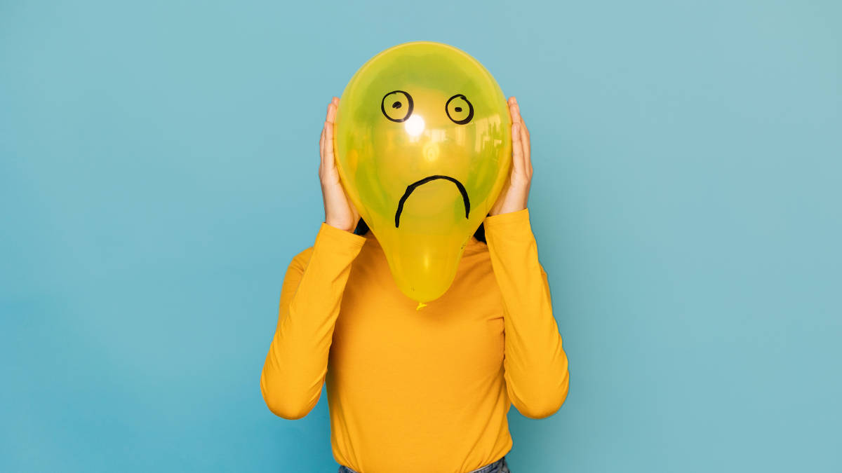 Eine Person im gelben Pullover hält sich einen gelben Luftballon vor das Gesicht, auf den ein trauriger Smiley gemalt ist.